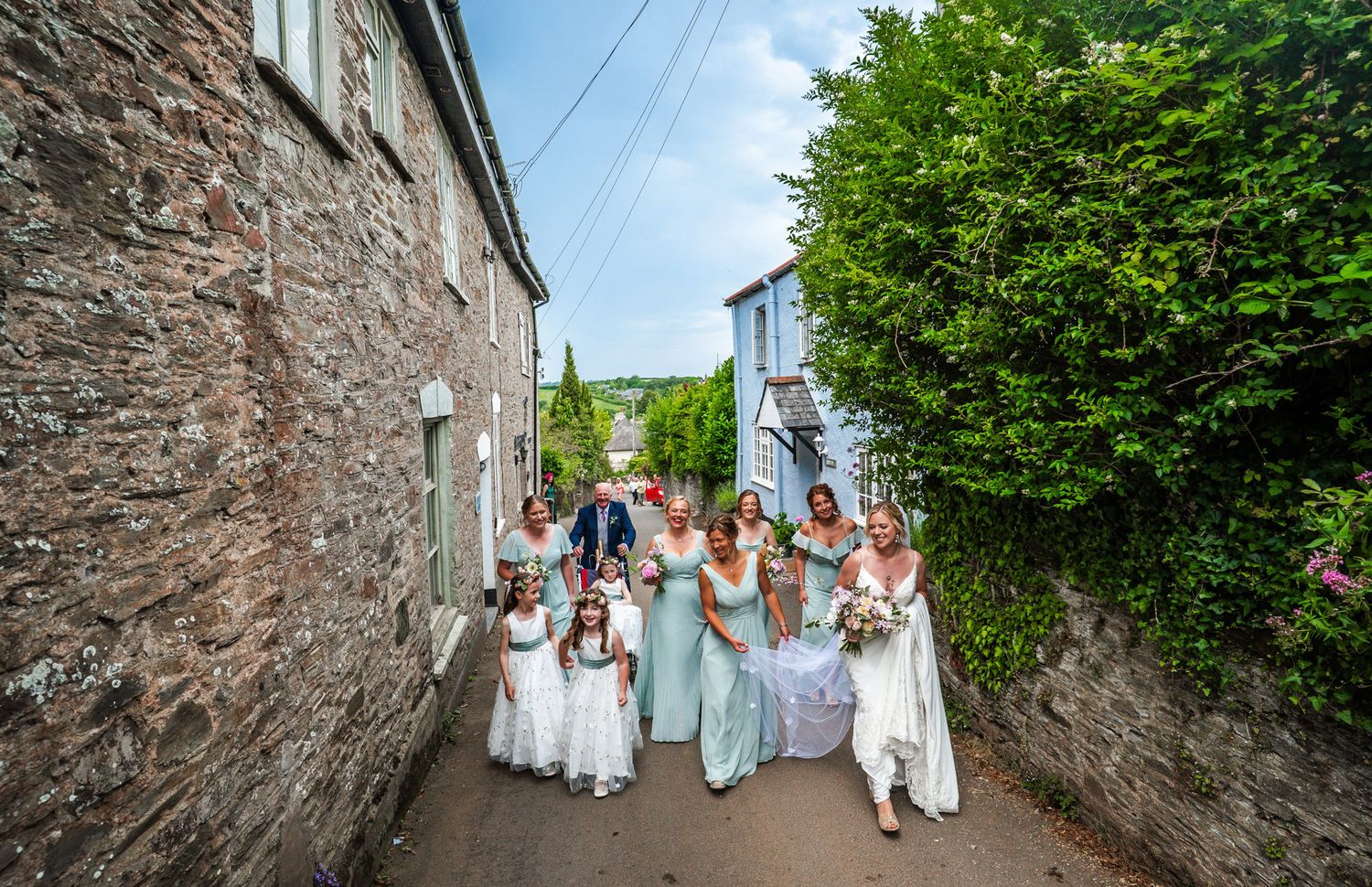 Wedding party walking along a rustic stone wall path surrounded by greenery and overhead power lines.