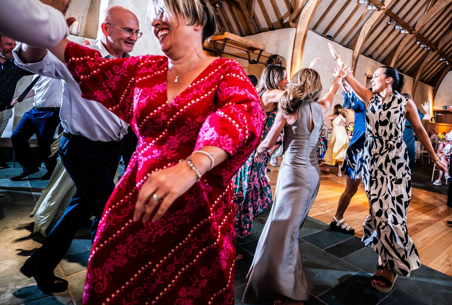 Wedding guests dance and celebrate on a wooden floor at a reception venue while wearing formal attire.