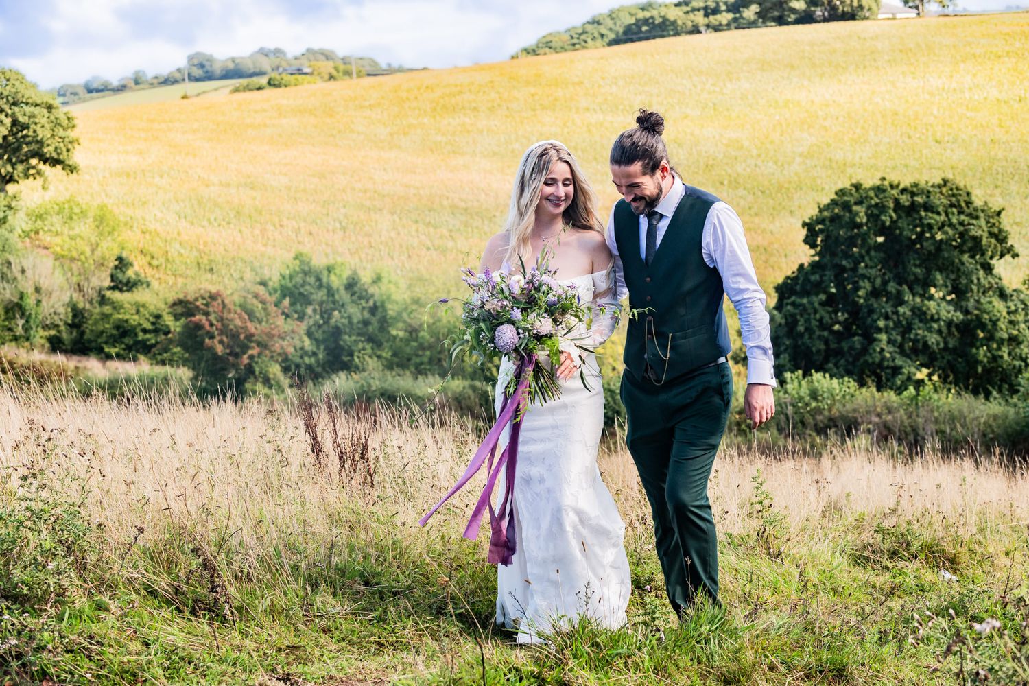 A couple walks through a golden summer meadow with rolling hills, holding a vibrant purple and white bouquet.