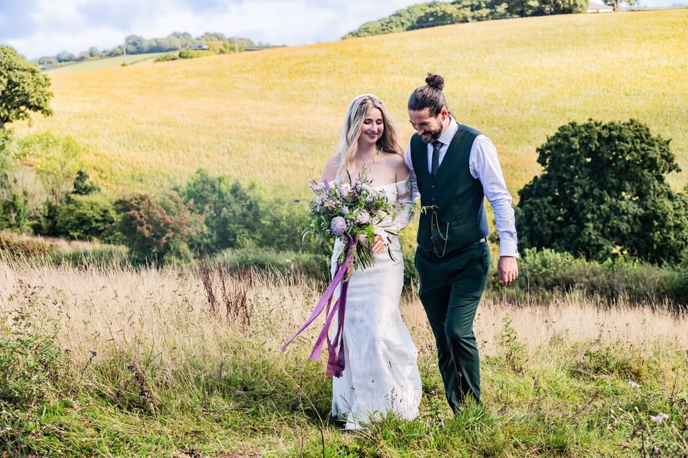 A couple walks through a golden summer meadow with rolling hills, holding a vibrant purple and white bouquet.