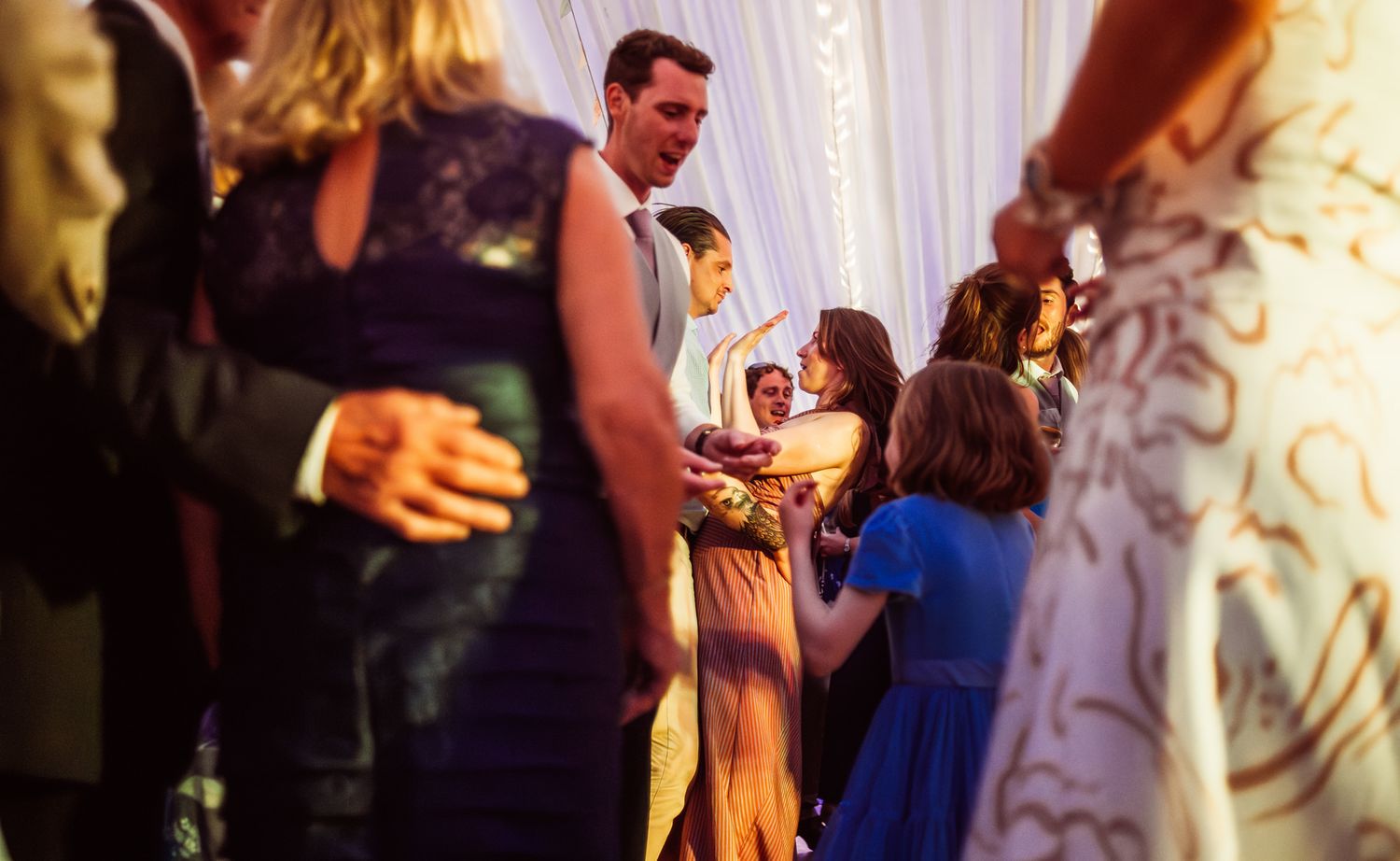 Wedding reception guests dancing and celebrating on a dance floor with decorative white curtains in the background.