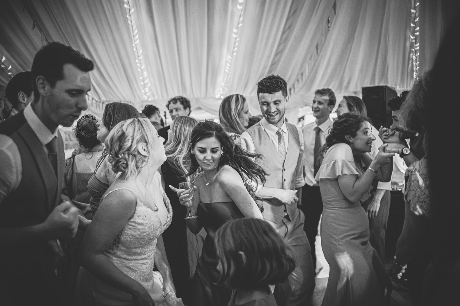 Wedding guests dance and celebrate on a crowded dance floor under dramatic lighting at an evening reception.