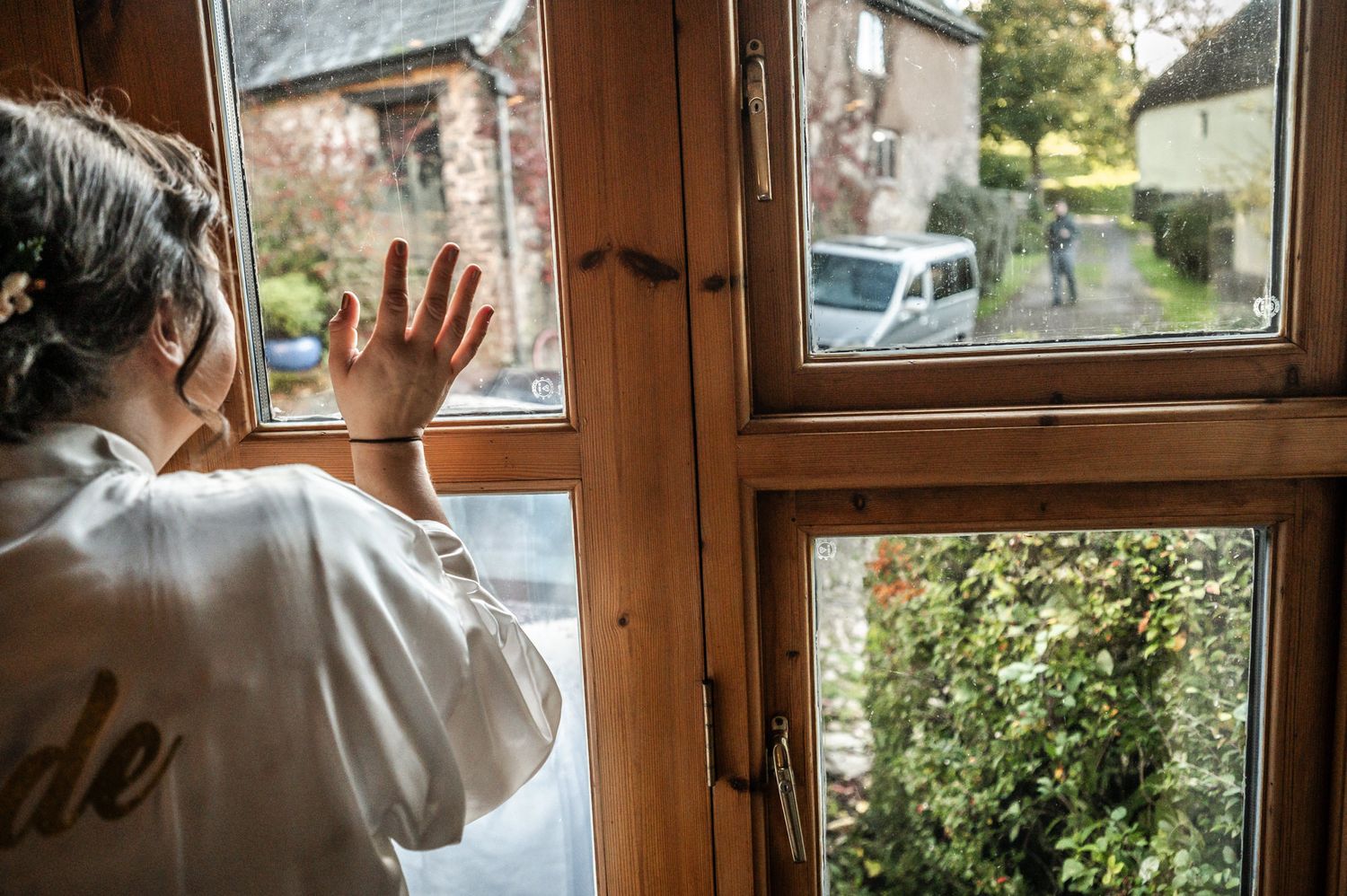 Someone in a white sweater looks through a wooden framed window at a car parked outside on a rural street.