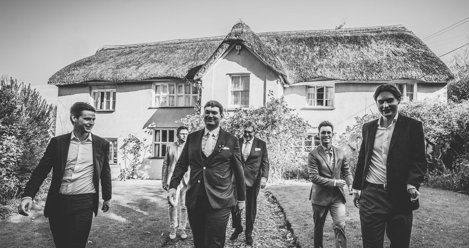 A black and white photo of people walking together outside traditional thatched roof cottages in a countryside setting.