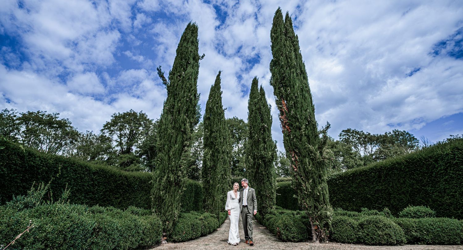 A couple stands between tall cypress trees in a manicured garden under dramatic blue skies.
