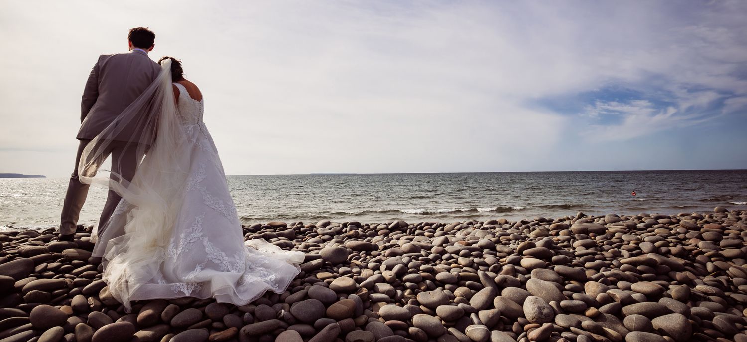 A scenic beach wedding ceremony setup with white chairs and floral arrangements along a rocky shoreline at sunset.