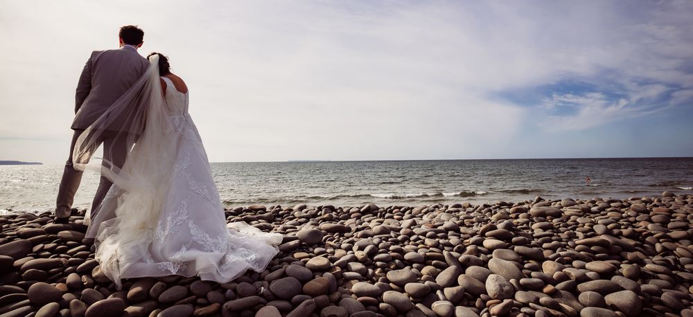 A scenic beach wedding ceremony setup with white chairs and floral arrangements along a rocky shoreline at sunset.