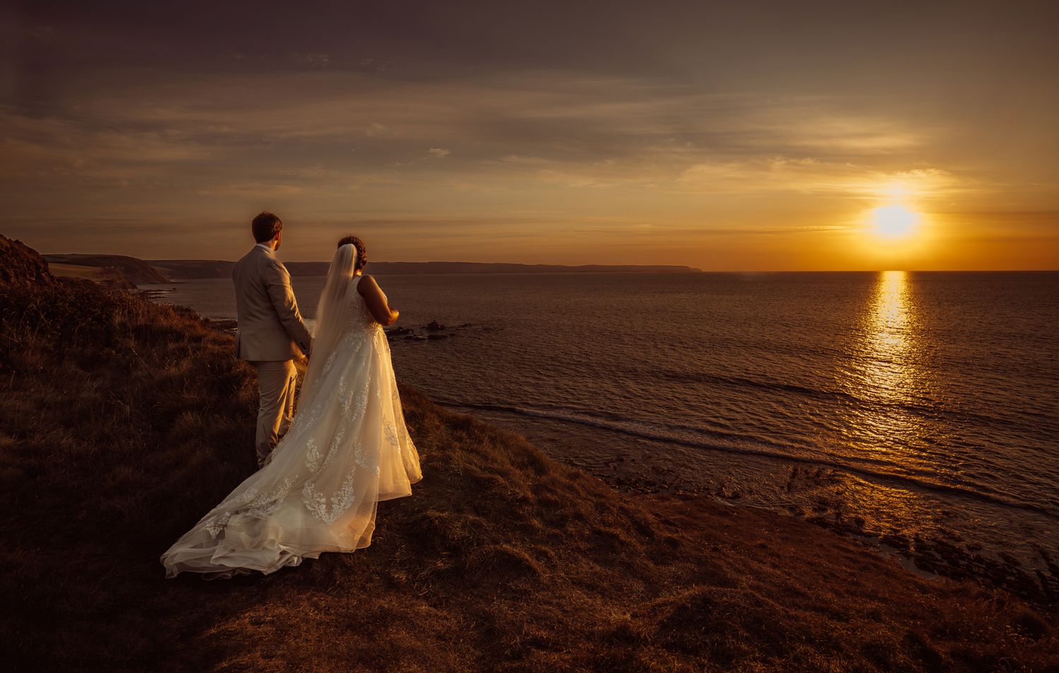 Silhouetted couple in wedding attire stands on coastal cliffs against dramatic orange sunset over ocean waves.