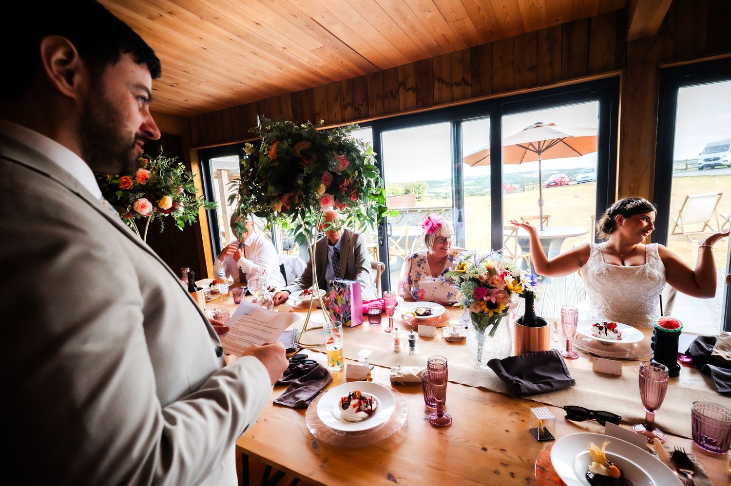 Guests gathered at a rustic restaurant table during a celebration with wine glasses and natural lighting through windows.