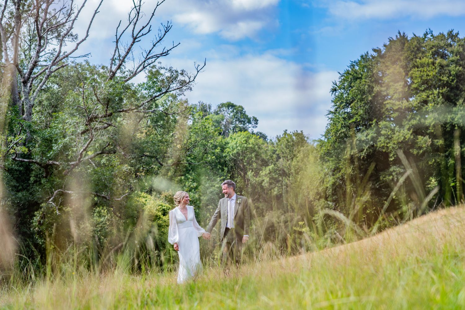 A couple walks together through a tall grass meadow on a sunny summer day with trees in the background.