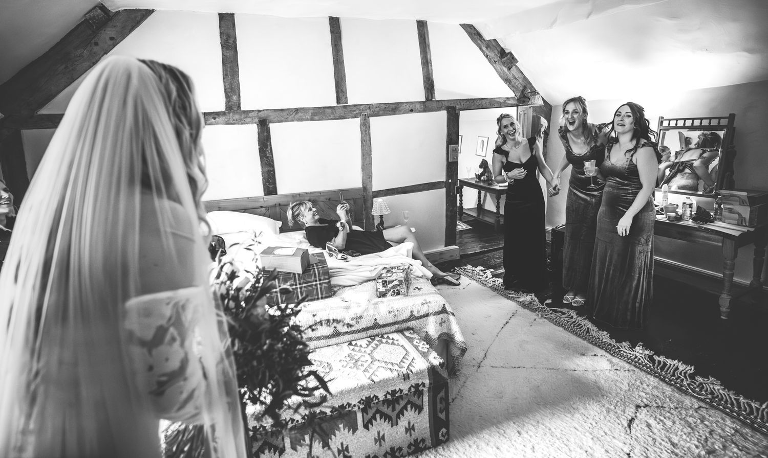 Black and white photo of bridesmaids watching bride get ready in rustic room with exposed timber beams.