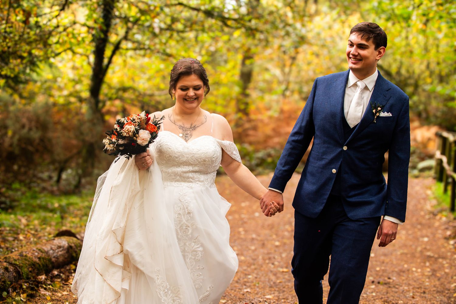 Newlywed couple walks hand in hand through a vibrant autumn forest path with golden leaves and natural light.