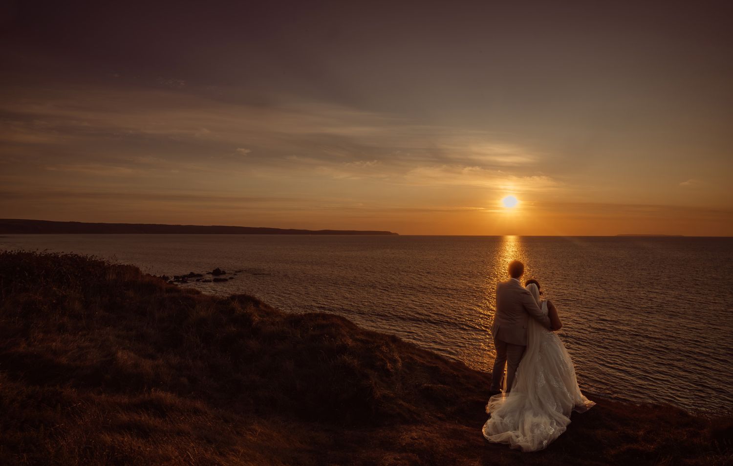 Silhouetted couple embraces on coastal cliffs during golden sunset over ocean horizon.
