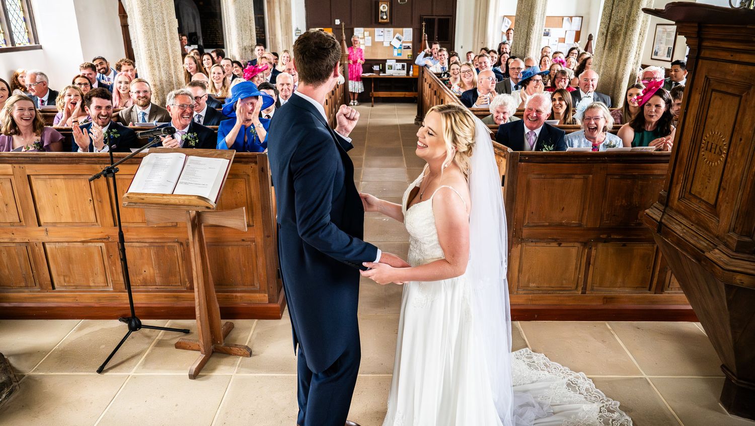 A wedding ceremony takes place in a beautiful stone church with wooden pews filled with guests watching the couple.