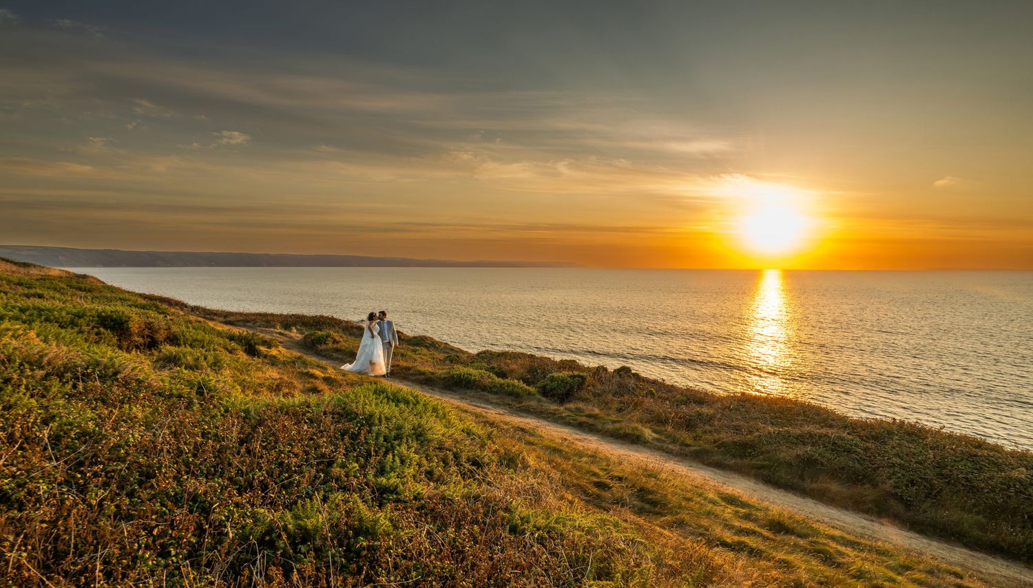 A dramatic sunset over the Pacific Ocean with golden light reflecting on the water and coastal cliffs in view.
