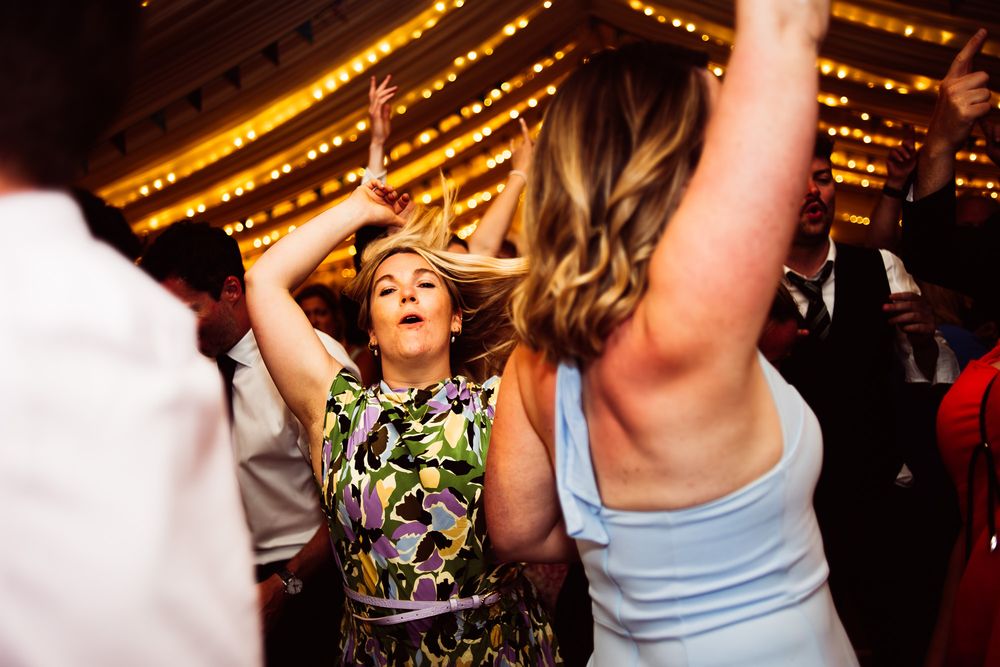 A lively crowd dances and celebrates on a dance floor under warm lighting at what appears to be a wedding reception.
