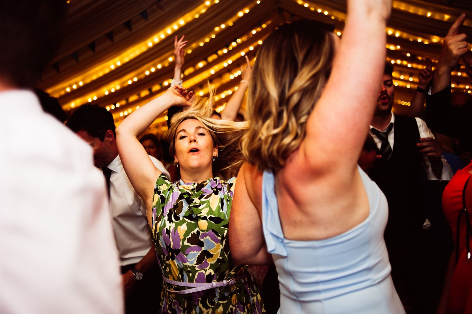 A lively crowd dances and celebrates on a dance floor under warm lighting at what appears to be a wedding reception.