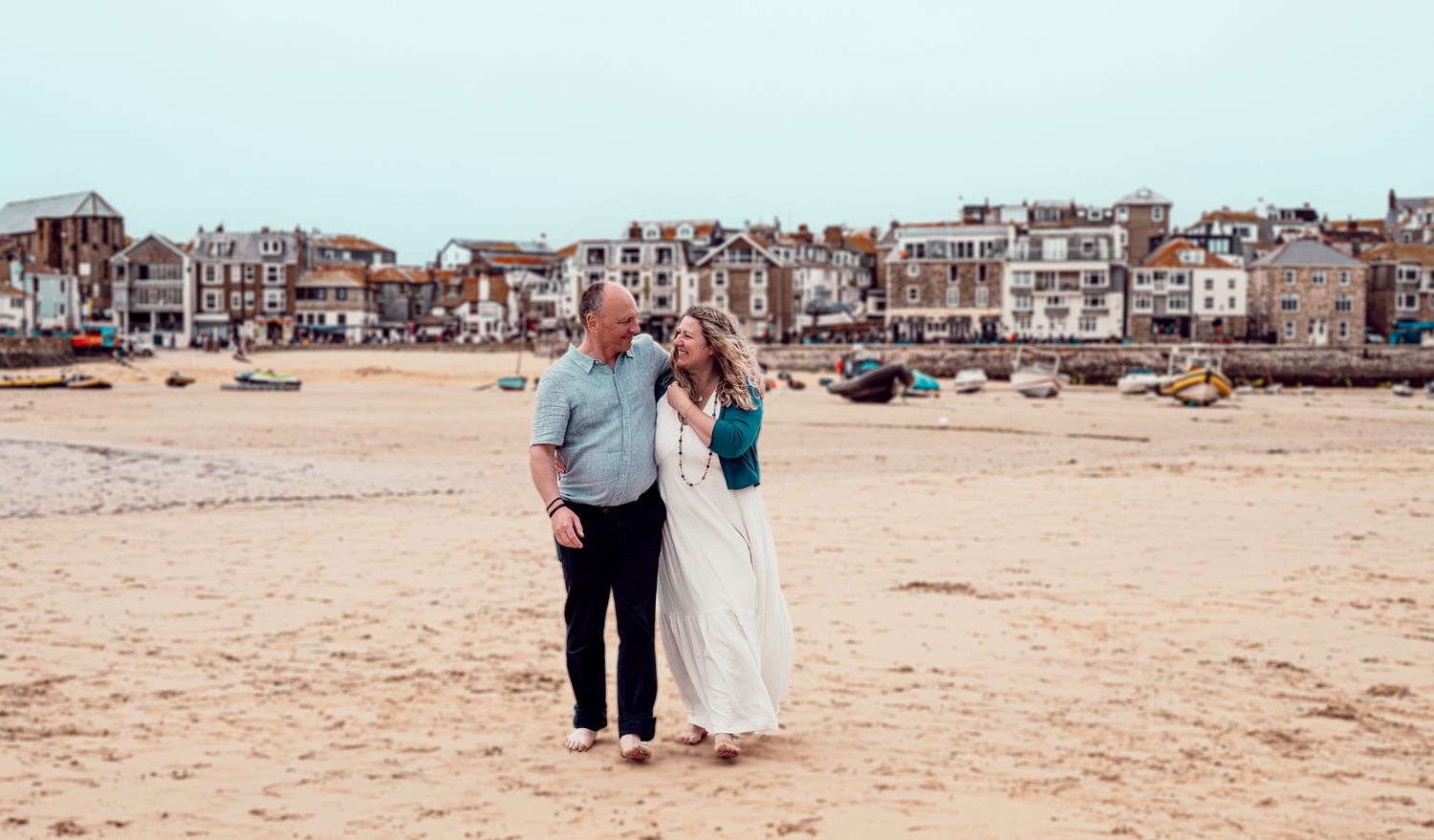 A couple walks together along a sandy beach with coastal buildings visible in the background.