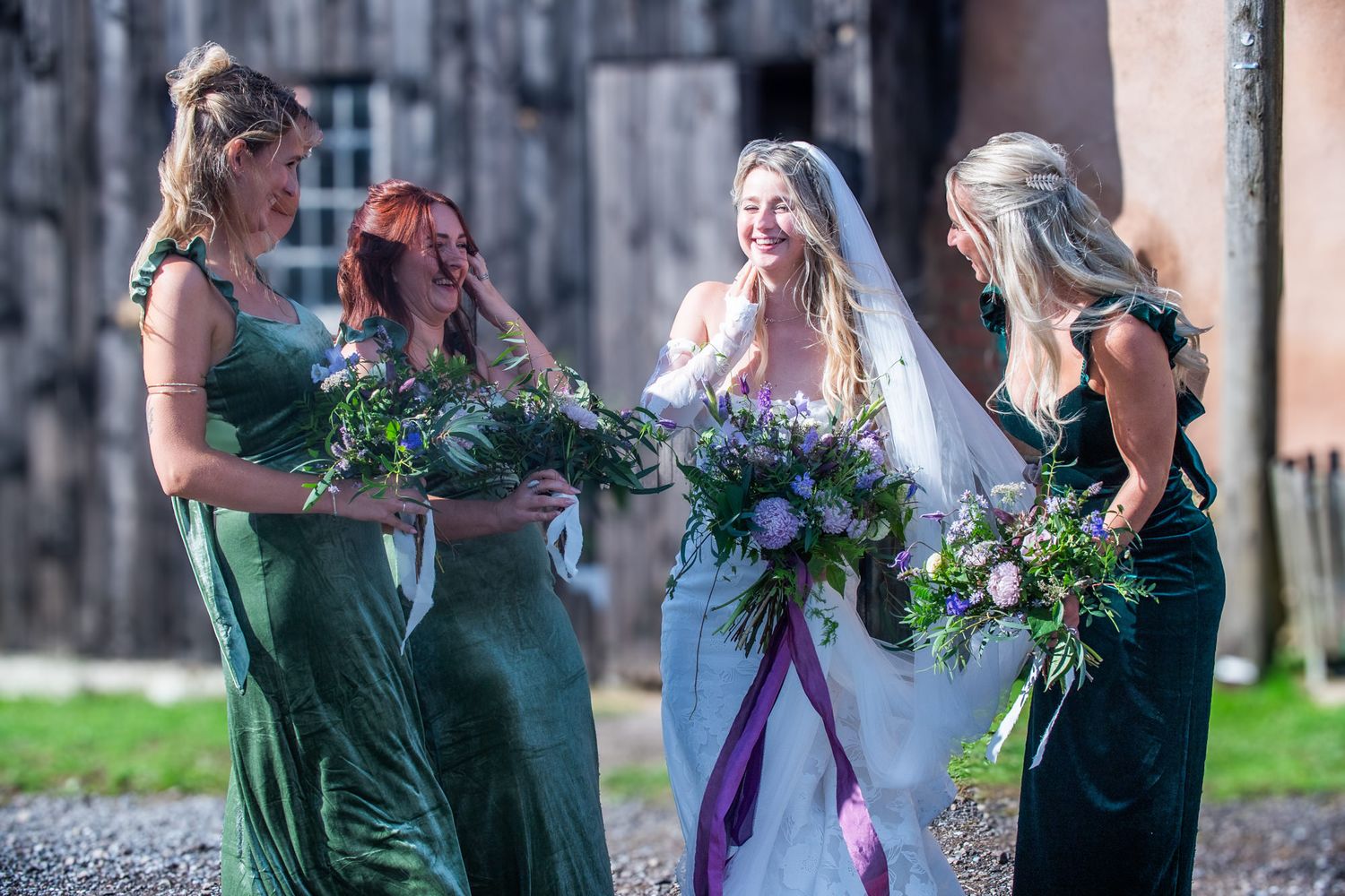 Bridesmaids in emerald green dresses share joyful moments with bride in white gown during outdoor wedding photo session.