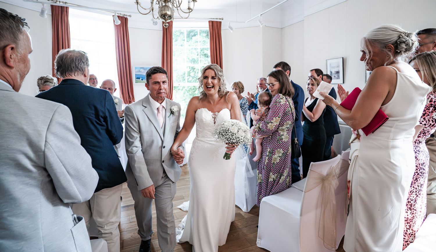 Wedding guests gather in a bright indoor venue with white walls and red curtains during a celebration.