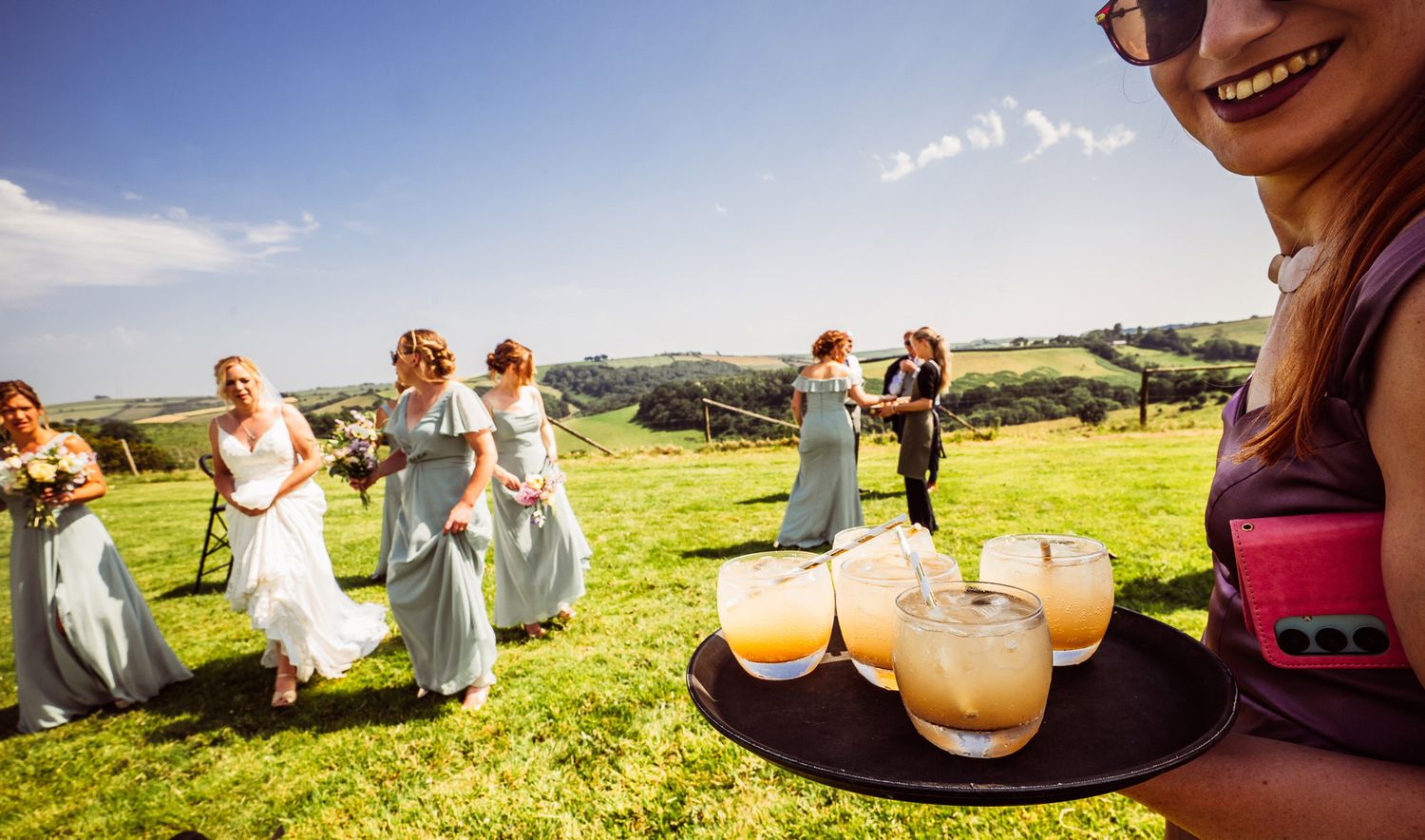 Wedding guests enjoy drinks and conversation while lounging on blankets in a sunny meadow with rolling hills.