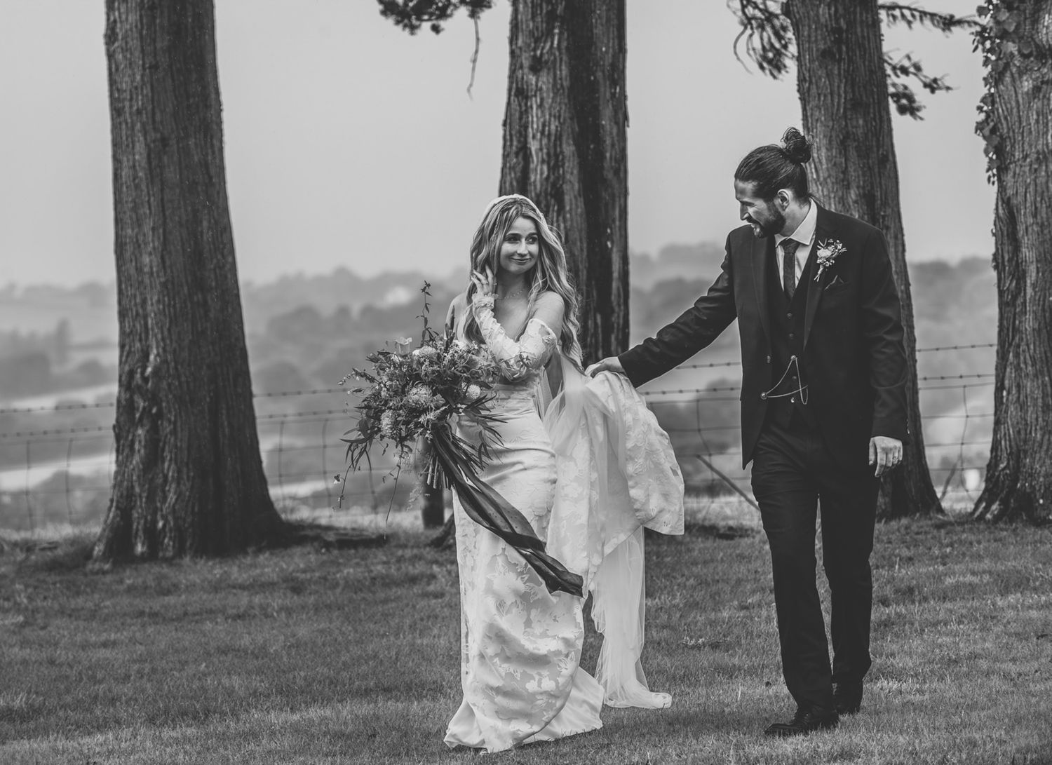 Black and white photo of a couple walking together through a scenic grove of tall pine trees.