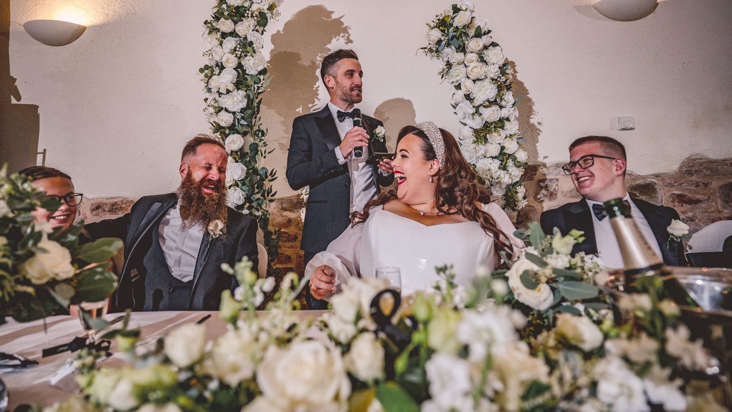 Wedding reception moment during a speech with guests seated at a decorated table with white floral arrangements.