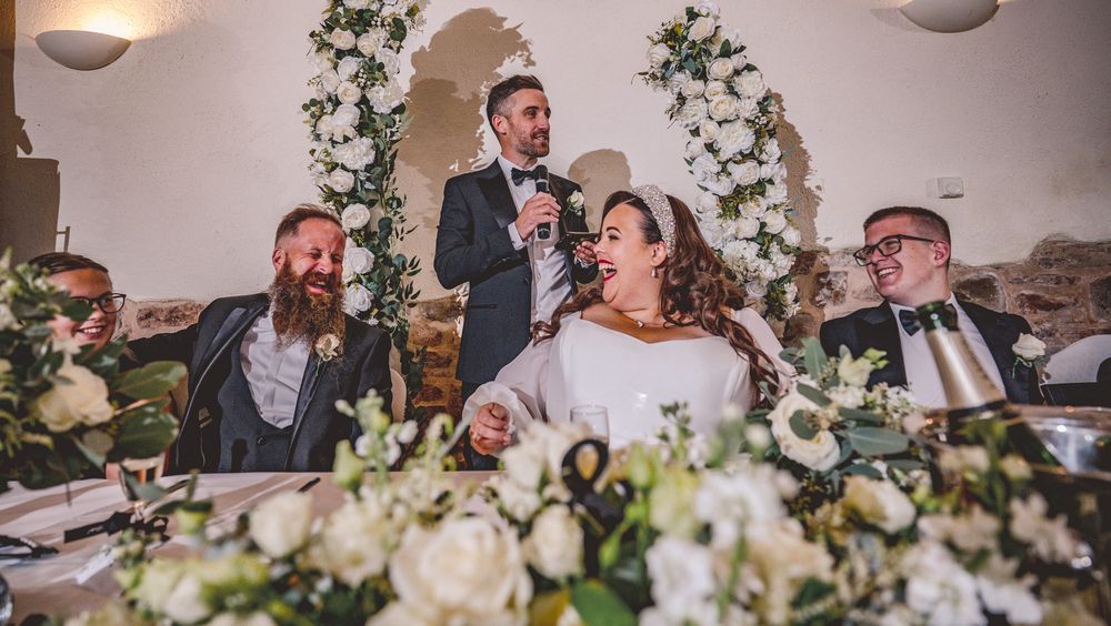Wedding reception moment during a speech with guests seated at a decorated table with white floral arrangements.