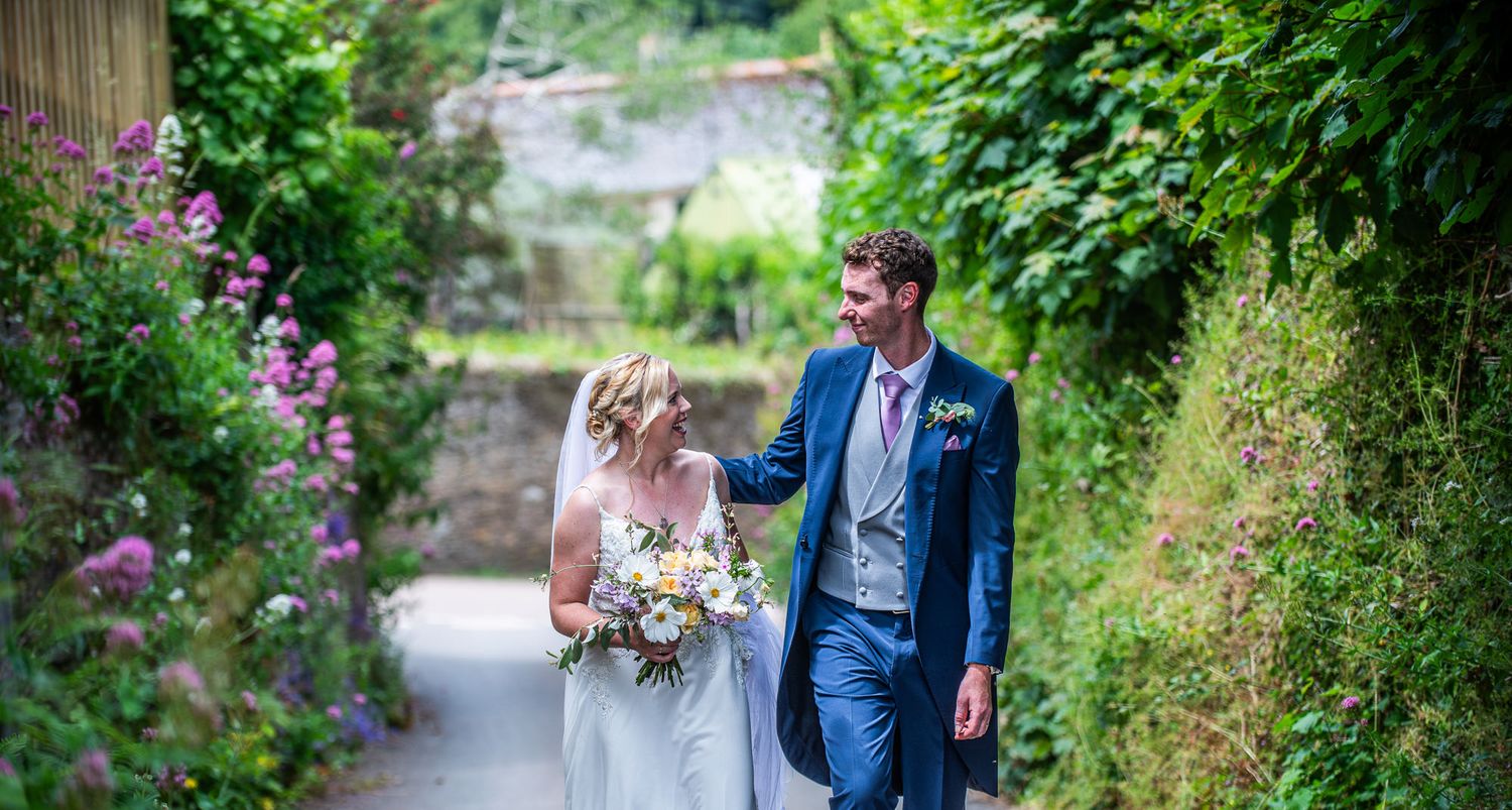 A couple walks together on a garden path surrounded by lush greenery and pink flowers on their wedding day.