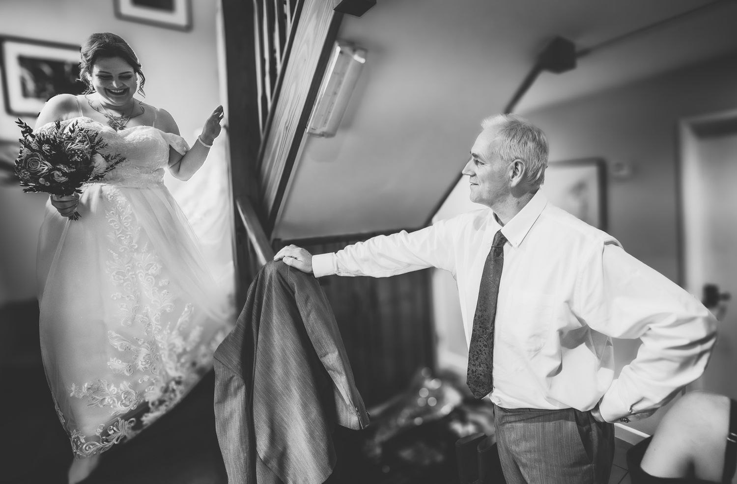 A black and white wedding photo showing a bride descending stairs while holding flowers as someone waits below.