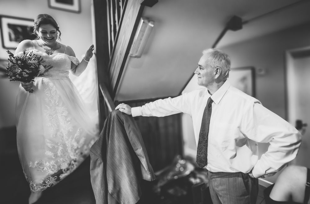 A black and white wedding photo showing a bride descending stairs while holding flowers as someone waits below.