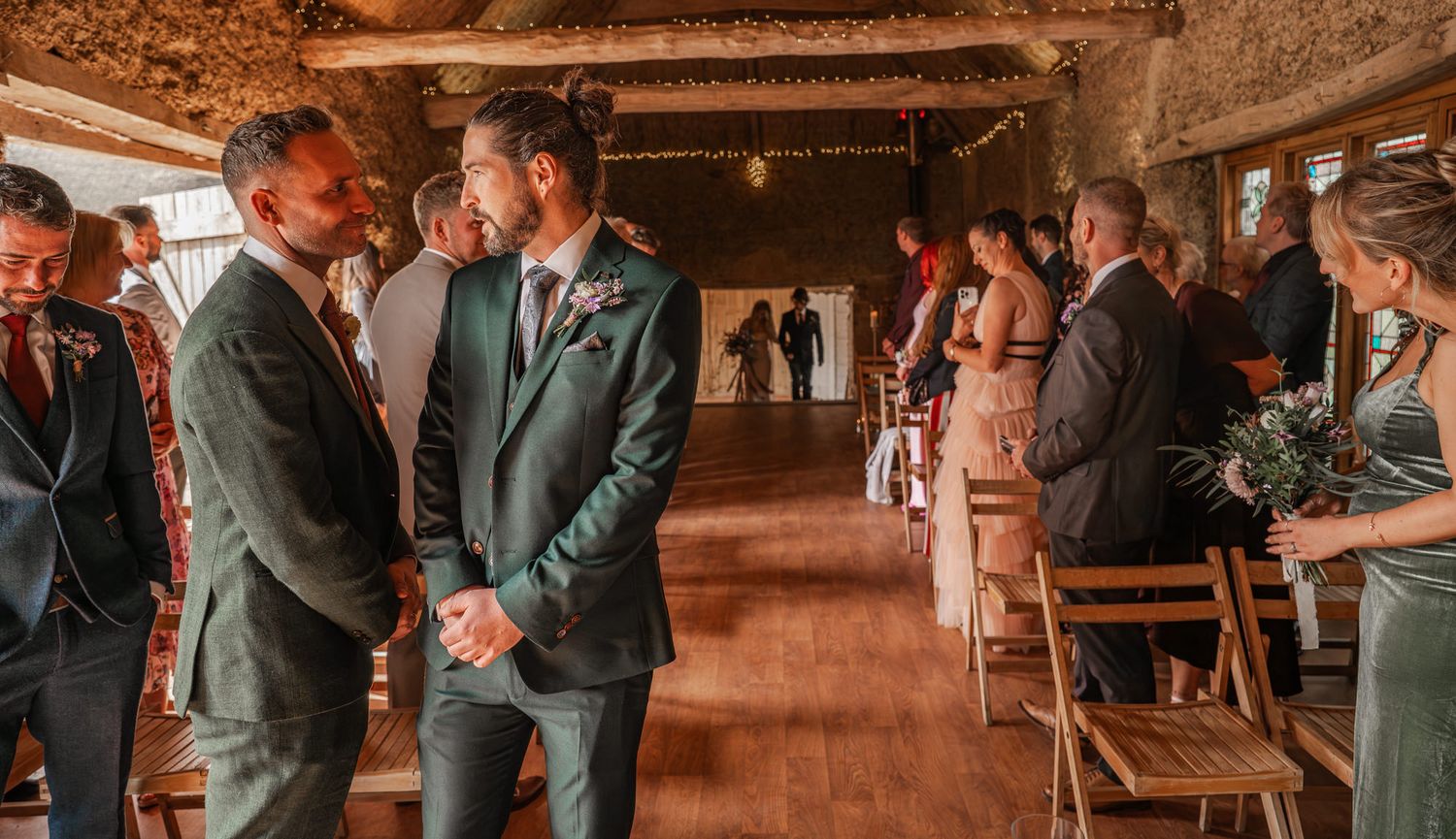 Two people in green suits share an intimate moment during a rustic barn wedding ceremony with guests seated nearby.