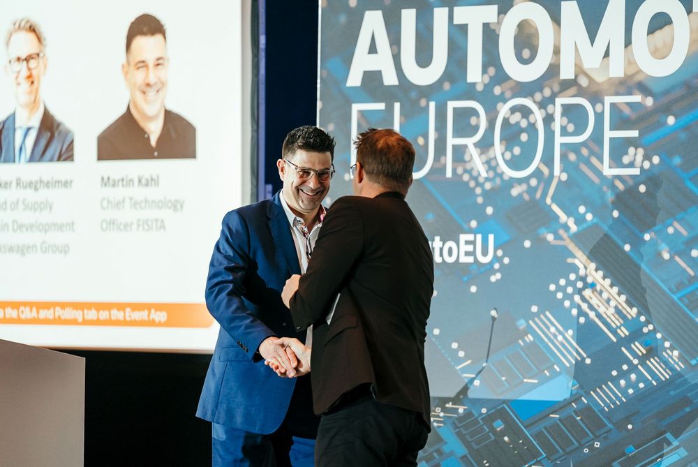Business professionals shake hands during a presentation with digital displays showing automotive industry content.
