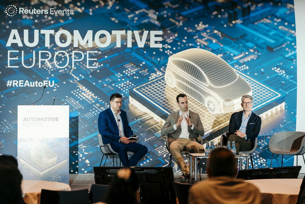 Business professionals seated on a panel at an automotive industry conference with blue lighting and backdrop.