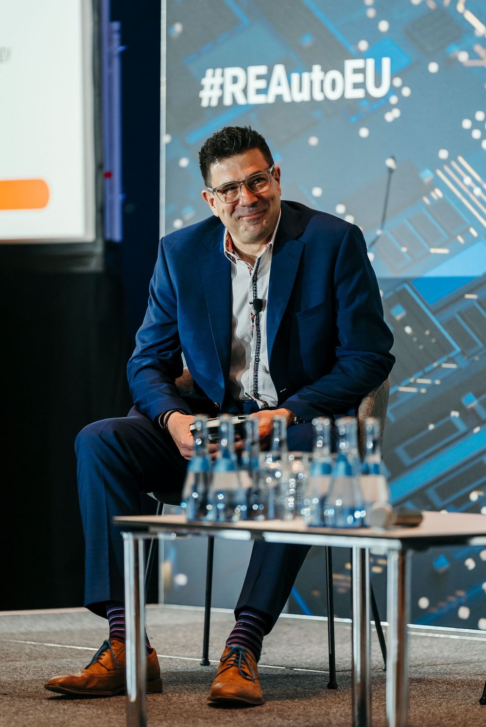 Professional in a navy suit sits on stage with crossed legs during a business conference presentation.
