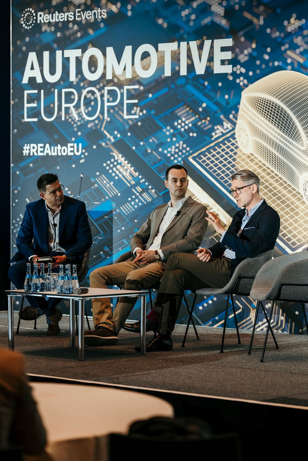 Panel discussion at Reuters Automotive Europe conference with speakers seated on stage against blue backdrop with circuit design.