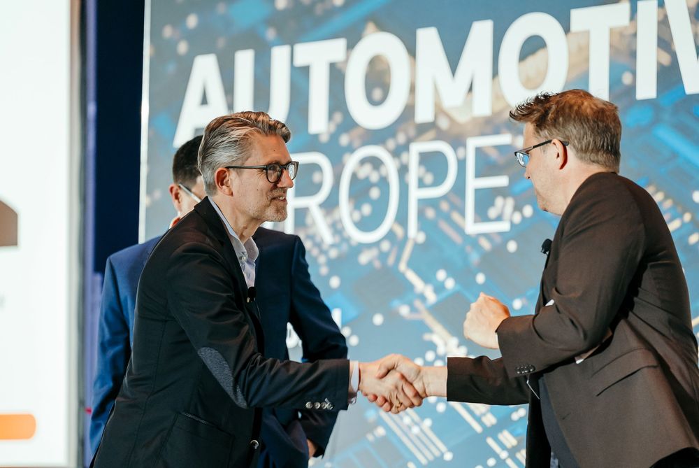 Two professionals in suits shake hands at an event with an Automotive backdrop display.