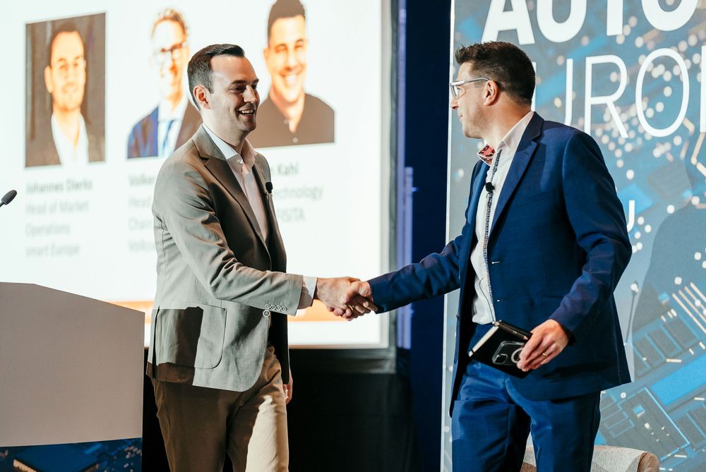 Business professionals shake hands during a corporate event with Auto signage visible in the background.