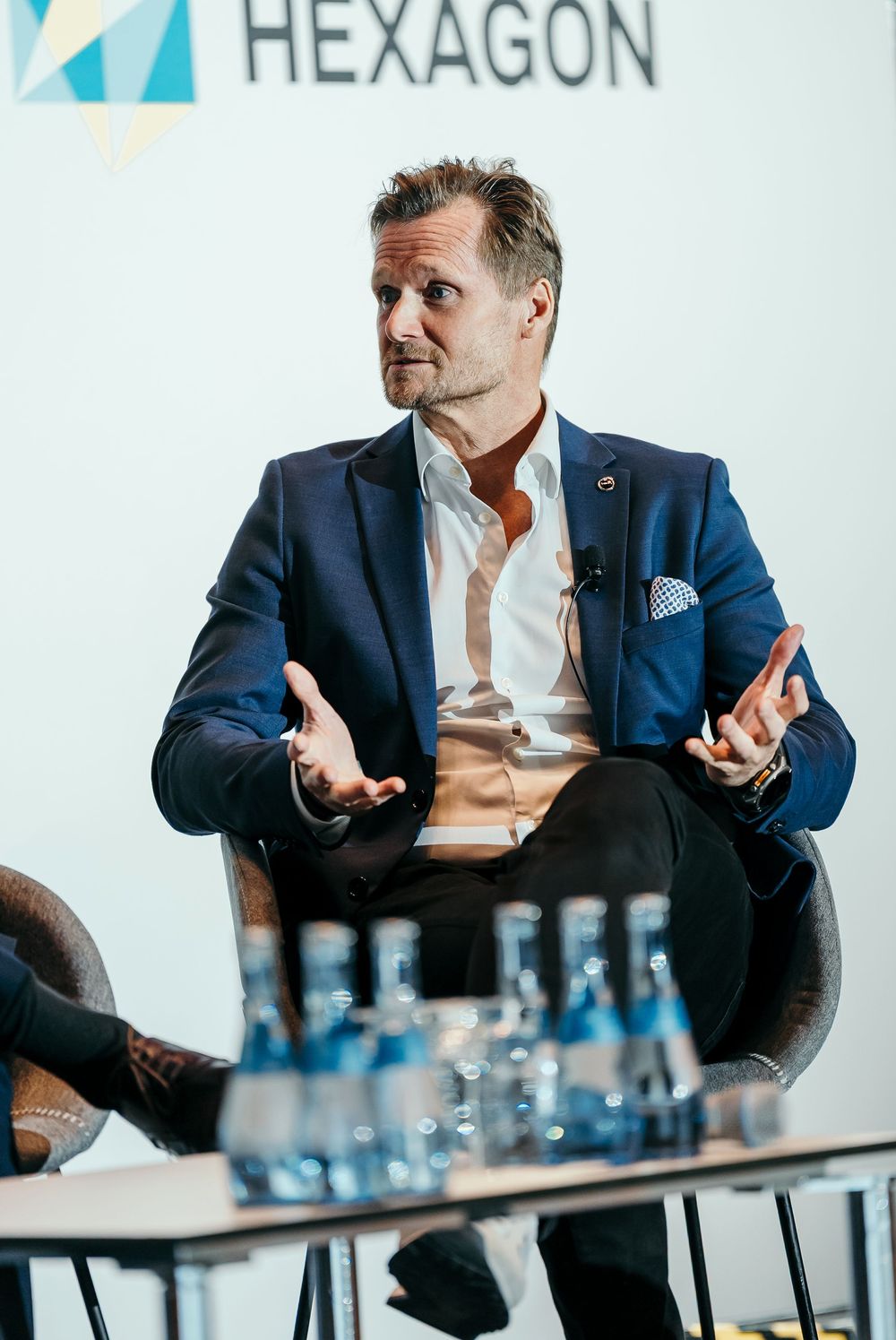Professional in a navy suit gestures expressively during a business meeting with water glasses on the table.