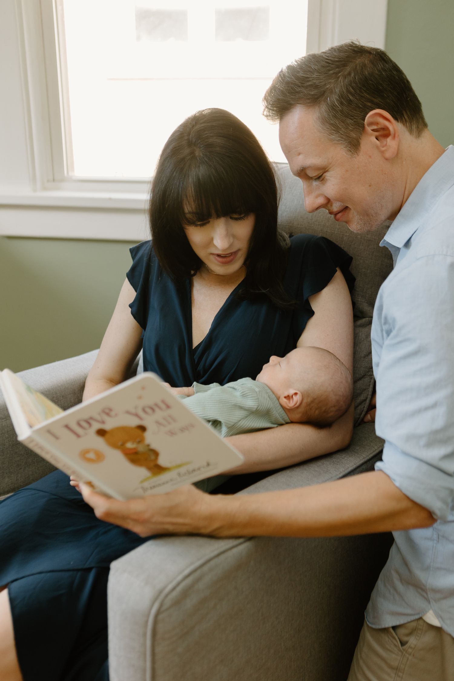 Family reading book together on grey couch in sunlit room.