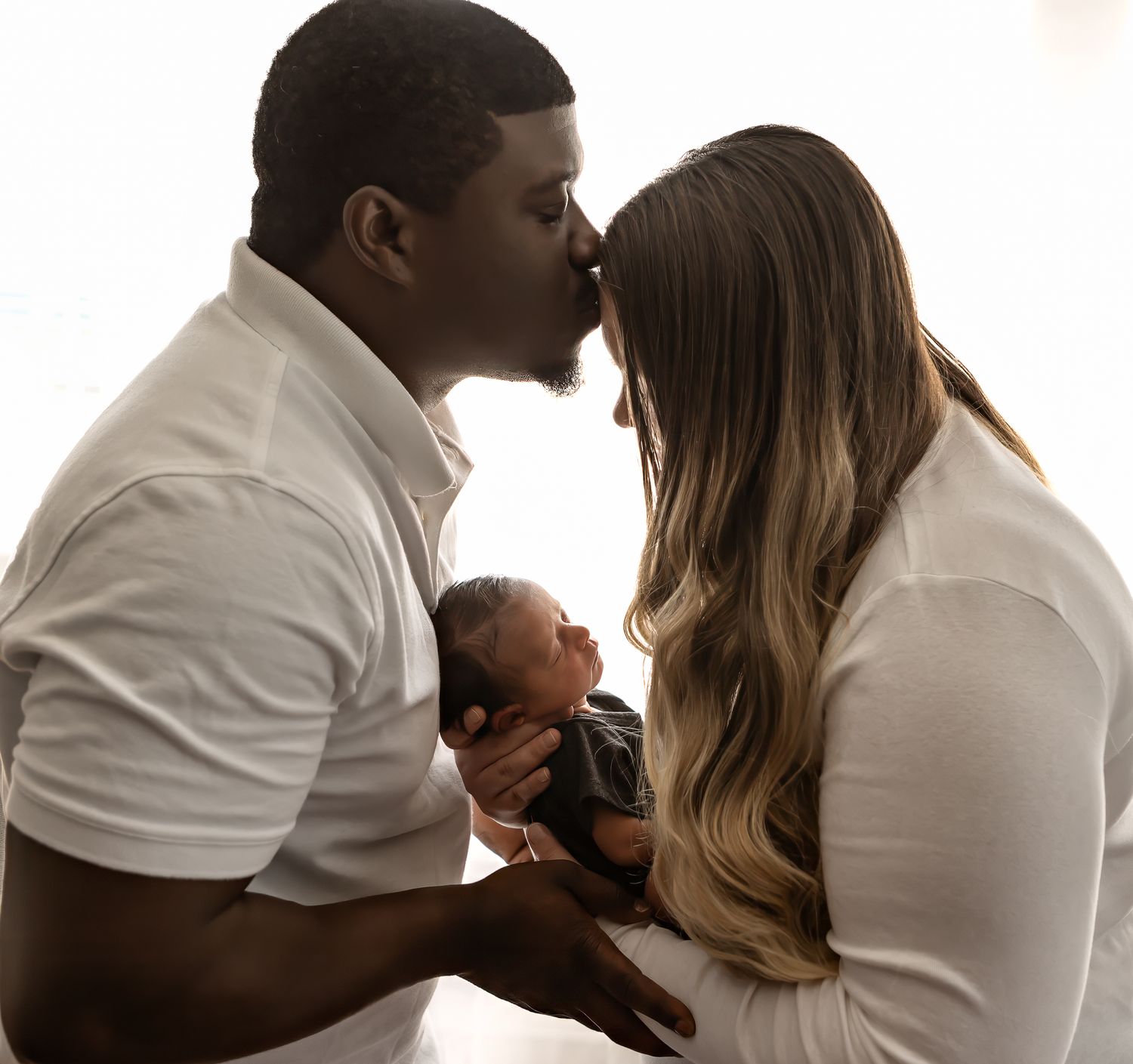 A tender family moment as parents share a kiss while holding their newborn baby between them.