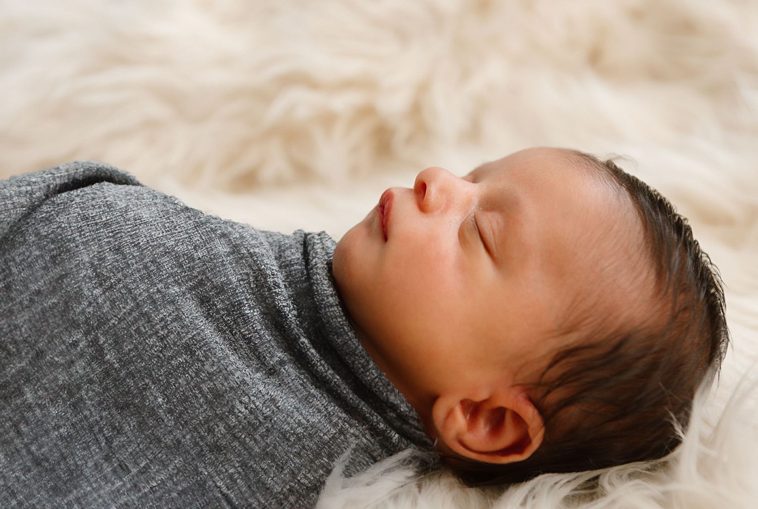 Peaceful newborn baby sleeping while wrapped in grey swaddle on soft furry surface.