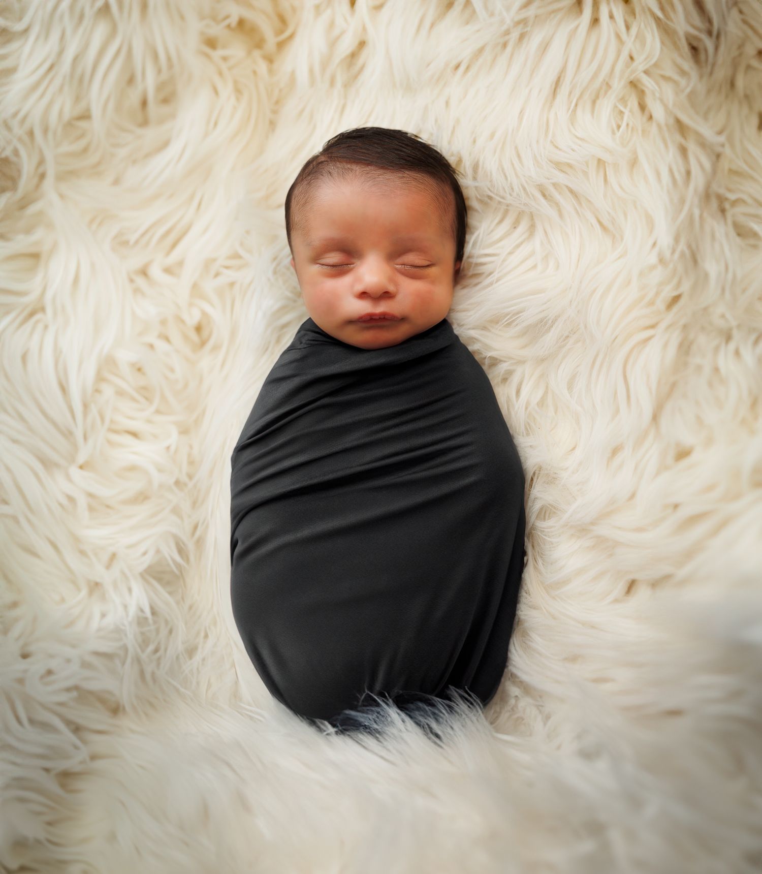 Sleeping newborn baby wrapped in black swaddle on white furry backdrop.