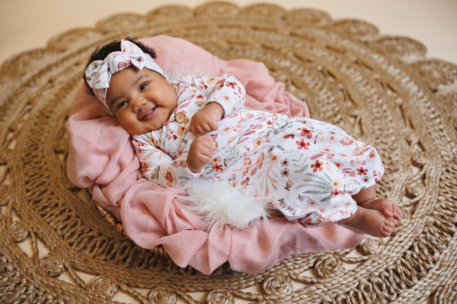 Baby wearing floral outfit and bow sleeping on pink blanket atop woven mat.