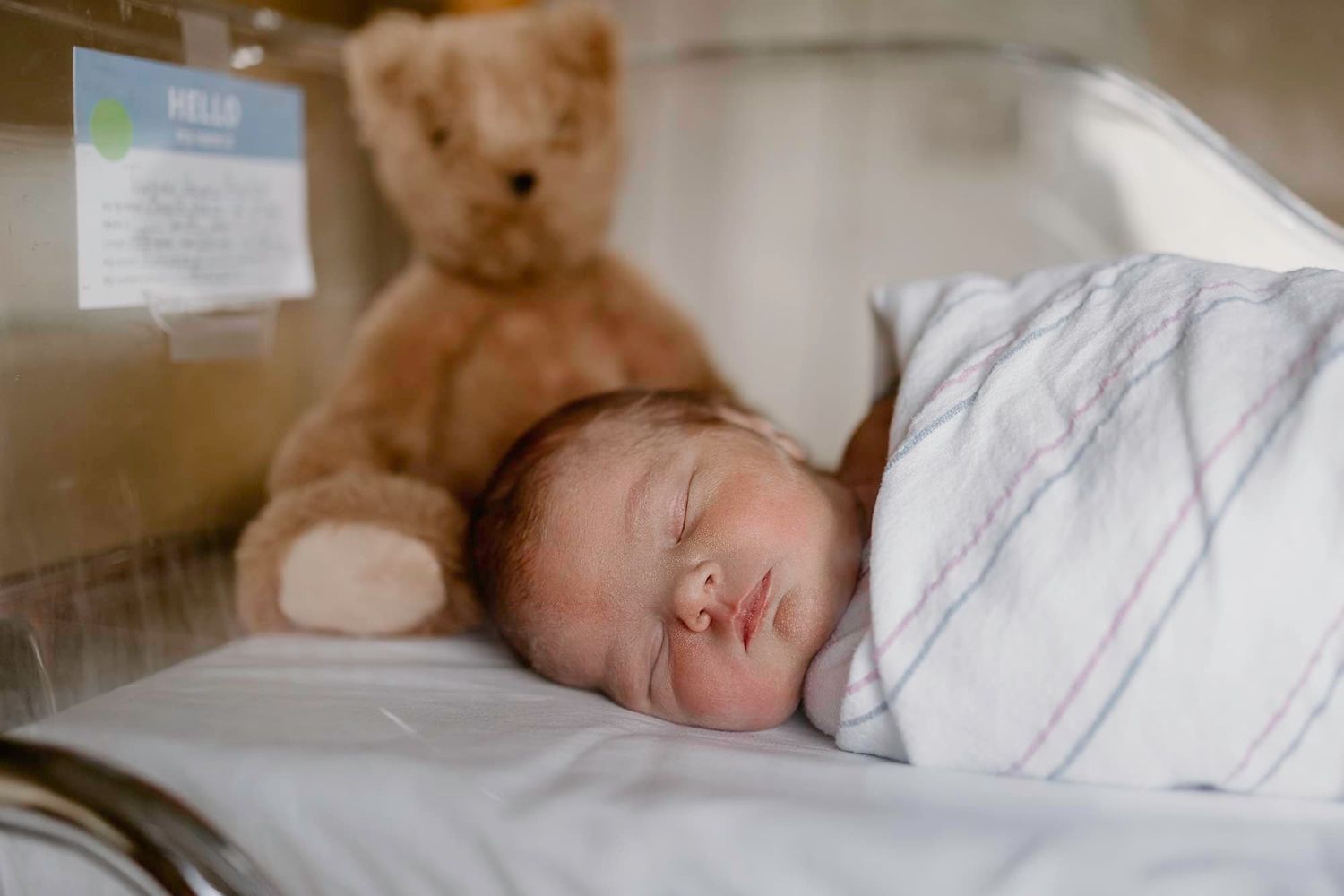 Sleeping baby in white outfit with brown teddy bear on striped bedding.