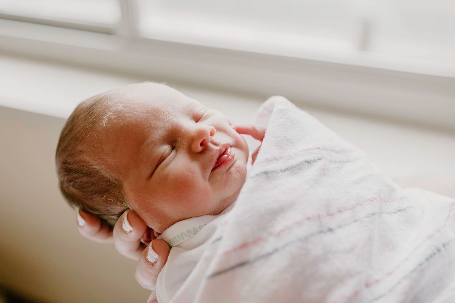 Close-up profile view of sleeping newborn in white swaddle against bright window light.