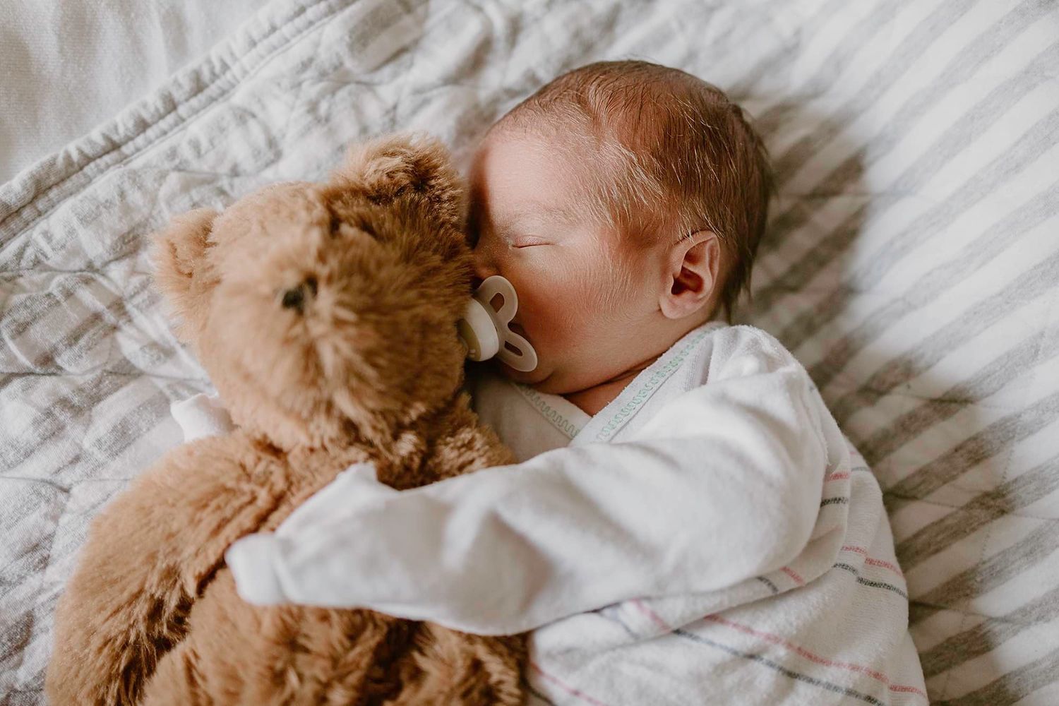 Peaceful newborn sleeping next to soft brown teddy bear on striped bedding.
