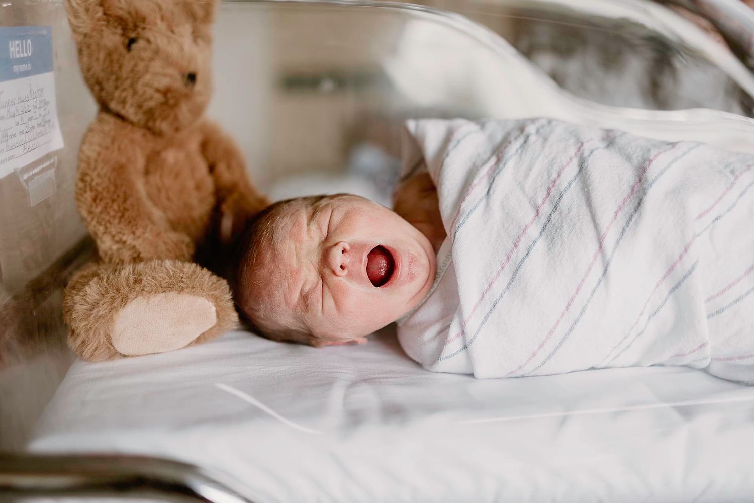 Baby sleeping peacefully in white swaddle with brown teddy bear beside them.