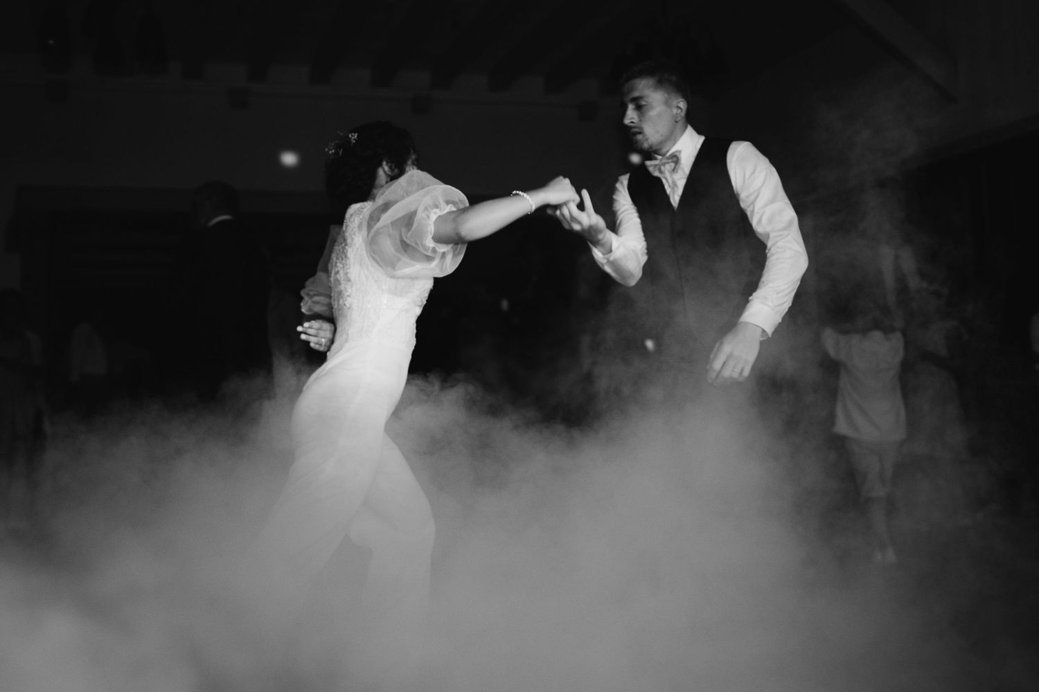 Black and white photo of bride and groom dancing together in dramatic fog on dance floor.