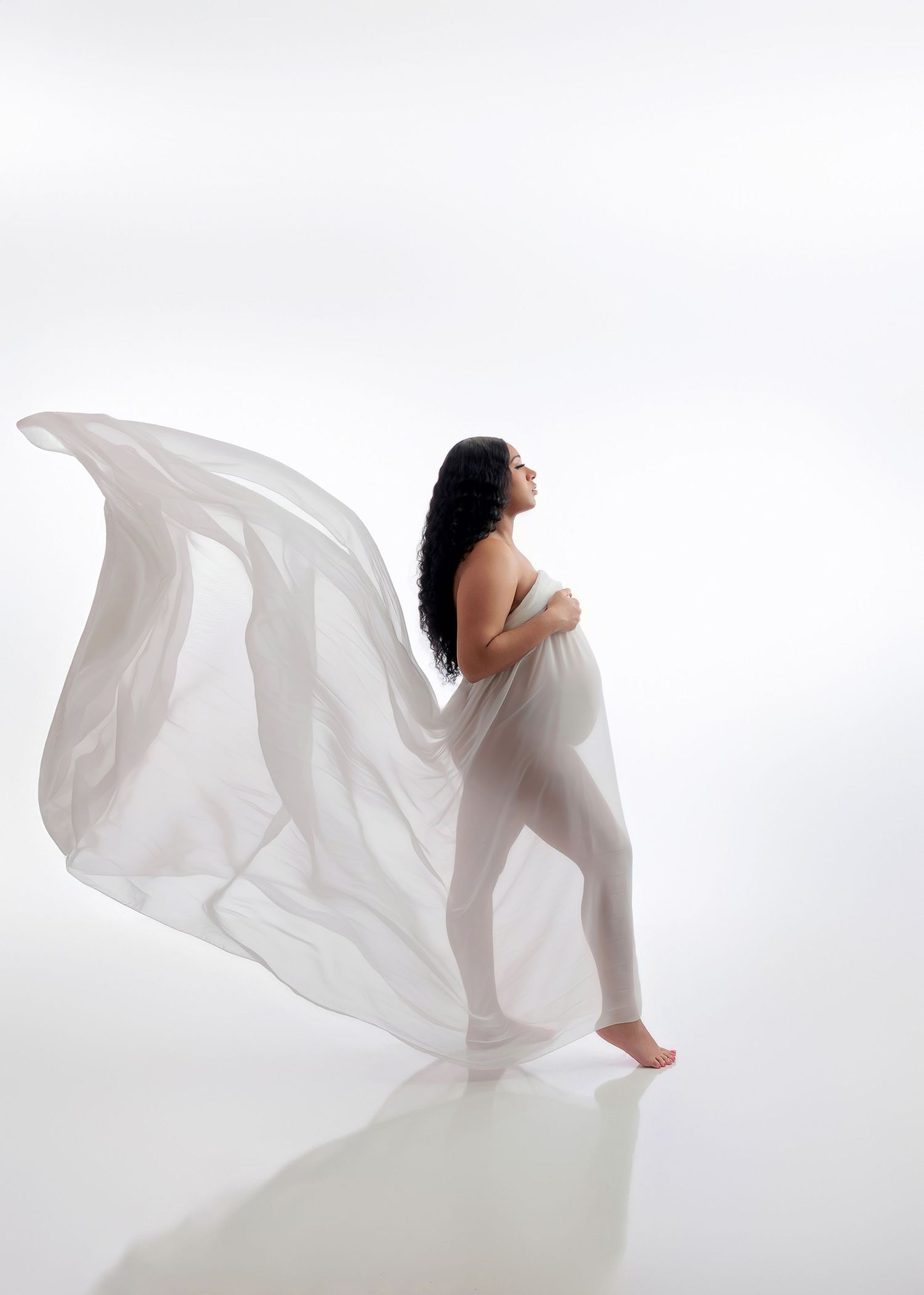 Pregnant woman holds billowing white sheer fabric as it flows behind her in white studio.